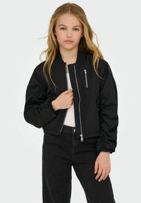 Giubbotto bomber nero con maniche a palloncino, chiusura anteriore con zip, una tasca con zip e polsini elastici. Indossato sopra una maglietta bianca e jeans neri.