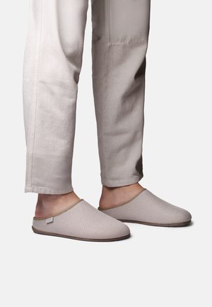 Jalad, millel on beežidest võrgust libisemiskindlad sussid, mida kannavad heledate beežide pükstega, ühevärvilise tausta ees.