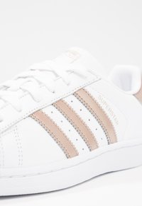 Sneaker bianca con tomaia in pelle liscia, caratterizzata da tre strisce in suede beige, perforazioni e suola in gomma testurizzata. Logo "SUPERSTAR" sul lato.