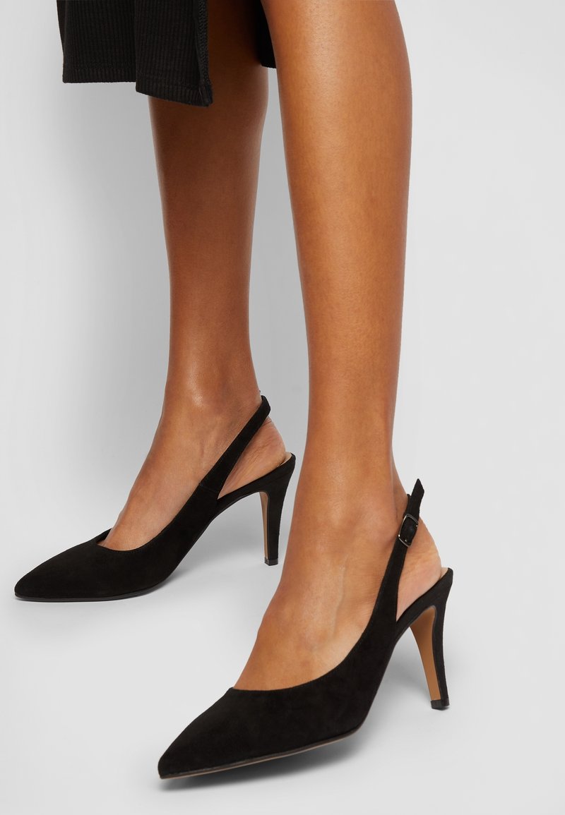 Jambes portant des escarpins slingback noirs en daim à bout pointu avec des talons fins de hauteur moyenne sur un fond neutre.