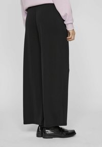 Crne široke hlače s elastičnom trakicom u struku, izrađene od mekog materijala, u paru sa sjajnim crnim mokasinkama, pokazujući glatku teksturu i čiste linije.