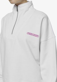 Sudadera de poliéster blanca con medio cierre en el cuello, que presenta un logo 'PEGADOR' en color rosa en el pecho. Tejido texturizado con un corte relajado y detalles de costura.