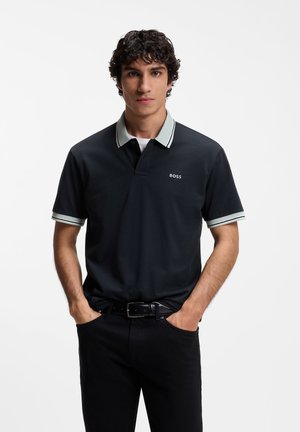 Junger Mann mit lockigem Haar, der ein schwarzes Poloshirt mit hellgrauem Kragen und "BOSS"-Logo trägt, Hände in den Taschen, vor weißem Hintergrund.