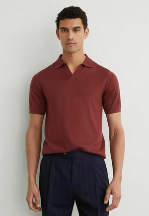 Mann trägt kurzärmeliges Poloshirt in Rostfarbe und dunkelblaue Nadelstreifenhose, steht vor einer hellgrauen Wand.