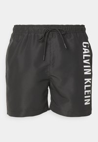 Crne kupaćice od glatkog materijala, s elastičnim pojasom koji se može zategnuti i bijelim natpisom "CALVIN KLEIN" sa strane.
