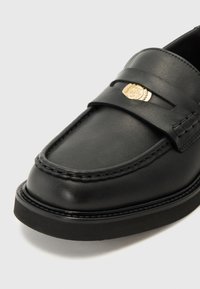 Black leather loafers featuring a gold emblem on the vamp, decorative stitching, and a flat rubber sole. Smooth texture with a polished finish.