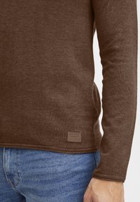 Brązowa koszula z długim rękawem wykonana z teksturowanej tkaniny, z małą metką przy szwie. Połączona z jasnoniebieskimi dżinsami.