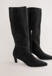 Schwarze kniehohe Stiefel aus Wildleder mit spitz zulaufender Zehenpartie, niedrigem Absatz und Reißverschluss an der Innenseite. Glatte Textur mit Nahtdetails.