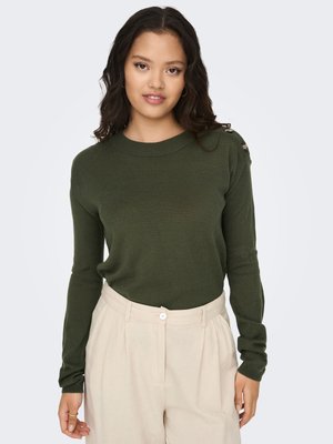 Mujer joven con cabello oscuro y ondulado, vistiendo un suéter verde oscuro y pantalones beige de talle alto, de pie frente a un fondo claro y liso.