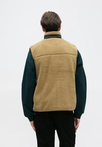 Chaleco de forro polar beige con cuello alto, con una superficie texturizada y ribetes negros. Lleva puesto sobre una camiseta de manga larga verde y pantalones negros.