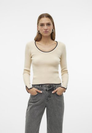 Top a maniche lunghe a coste di colore crema con scollatura ampia e bordi neri, abbinato a jeans grigi a vita alta. Tessuto morbido, silhouette aderente.