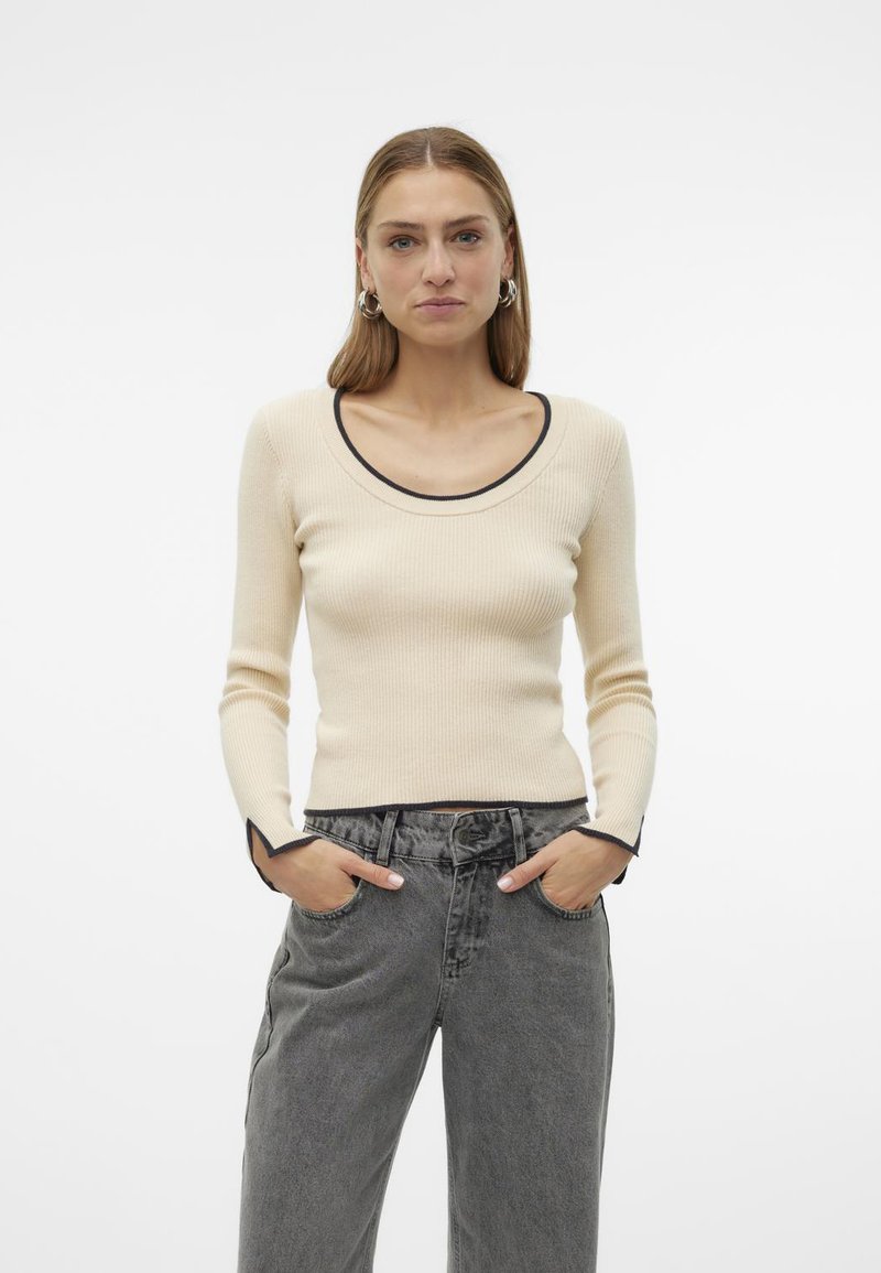 Top a maniche lunghe a coste di colore crema con scollatura ampia e bordi neri, abbinato a jeans grigi a vita alta. Tessuto morbido, silhouette aderente.