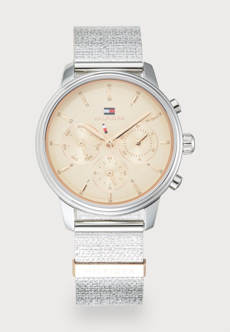 Montre-bracelet en métal argenté avec un cadran doré clair, trois sous-cadrans et un bracelet argenté texturé présentant un accent doré avec le nom de la marque.