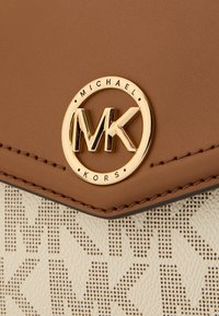 Karamelli nahkakott, millel on kuldne MK-logo, tekstureeritud kreemivalge taustaga, millel on peen MK-muster.
