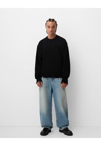 Črn oversized pleten pulover z rebrastimi manšetami, nosi se z svetlo modrimi širokimi hlačami z bledim prehodom in zvite robove. Črna obutev.