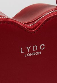 Rode leren handtas met een gegolfde bovenrand, zilveren ritsluiting en reliëflogo "LYDC LONDON" in zilver. Gladde textuur.