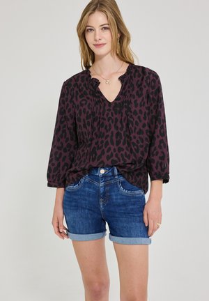 Ung kvinde med blondt hår iført en mørklilla og sort leopardmønstret bluse og blå denimshorts med opkrogede ben.