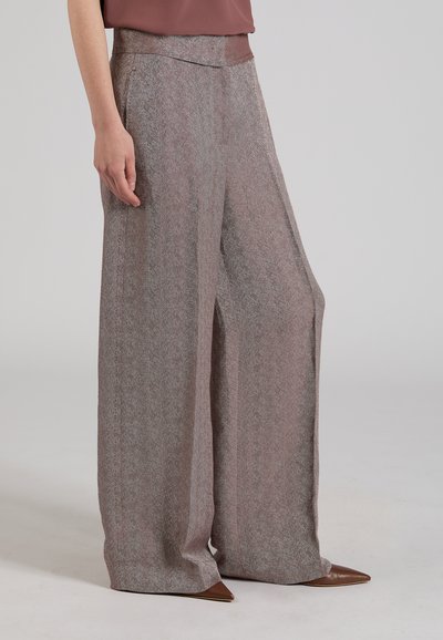 Donna che indossa pantaloni grigi a vita alta e gamba larga con trama e scarpe marroni con punta, con una maglia malva infilata, in piedi su uno sfondo semplice.