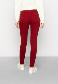 Červené skinny džíny s středně vysokým pasem, zadními kapsami a hladkým, elastickým materiálem. Nosené s bílými teniskami.