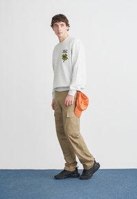Graues Sweatshirt mit grünem Kleeblatt-Design, khakifarbene Cargohosen, schwarze Turnschuhe und eine orangefarbene Kappe in der Hand. Einfaches, lässiges Design.