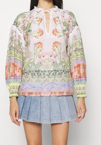 Blouse à motif floral, tissu léger, poignets élastiques et encolure en trou de serrure. Le design intègre diverses couleurs pastel et des détails complexes.
