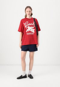 T-shirt gráfica vermelha com logo branco e design de cavalo, combinada com calções de ganga escuros, meias cinzentas e mocassins pretos, apresentando um corte descontraído.