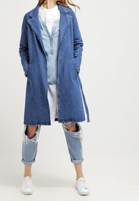 Dlouhý deninový kabát v tmavě modré barvě, se klopovým límcem, bočními kapsami a volným střihem, nošený přes světle modrý vícerozměrný outfit a tenisky.