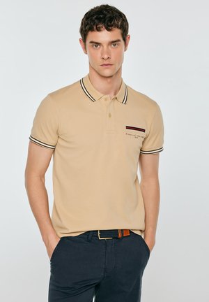 Młody mężczyzna w beżowej koszulce polo z paskowanym kołnierzykiem i rękawami, granatowe spodnie, stoi z rękami w kieszeniach na jednolitym tle.