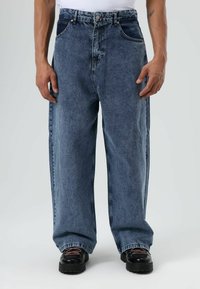 Blauwe spijkerbroek met een losse pasvorm, een zware textuur, vijf zakken en contrasterende zijpanelen. Gecombineerd met zwarte chunky schoenen.