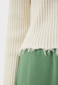 Pull côtelé crème avec ourlet effiloché, associé à un pantalon tricoté vert, présenté de la taille vers le bas sur un fond neutre.