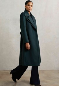 Naine, kes kannab pika tumeroheline jope laiade krae ja mustade laiad jalatsid ning mustade kontsaga saapadega, kõndimas ühtlase heleda tausta vastu.