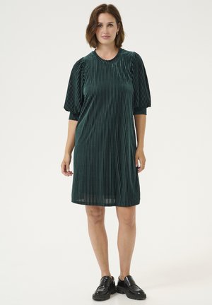 Grünes Samtkleid mit vertikalen Streifen, kurzen puffärmel, runder Ausschnitt und lockerer Passform. Getragen mit schwarzen Schuhen mit dicker Sohle.