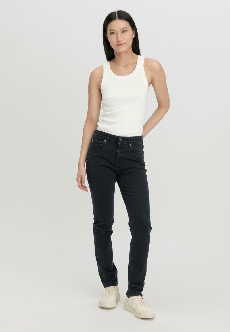 Schwarze Denim-Jeans mit fünf Taschen, Knopfverschluss und hellen Stickdetails. Kombiniert mit einem eng anliegenden weißen Tanktop und cremefarbenen Sneakers.