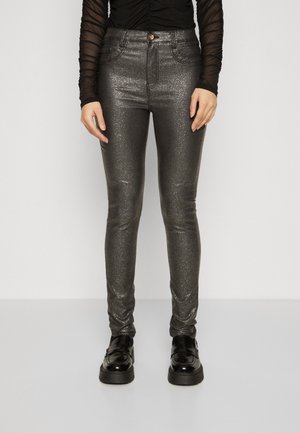 Kobieta ubrana w błyszczące, ciemnoszare, dopasowane spodnie, przezroczystą czarną bluzkę z długim rękawem oraz czarne loafersy na platformie, stojąca na białym tle.