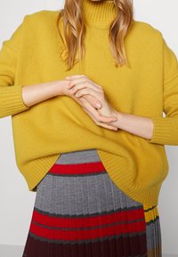 Osoba ubrana w musztardowy sweter z golfem i spódnicę plisowaną w poziome paski w kolorach czerwonym, szarym, żółtym i brązowym, trzymająca ręce złożone.