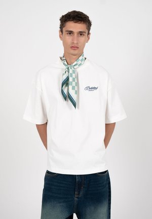 Młody mężczyzna ubrany w białą, oversize'ową koszulkę z napisem "Prohibited Racing Team" oraz zielono-białą kraciastą apaszkę na szyi, w połączeniu z wyblakłymi dżinsami.