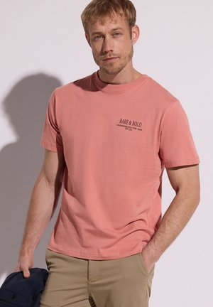 Homme portant un T-shirt rose saumon avec le texte « Bare & Bold », pantalon kaki, tenant une veste bleu marine, debout contre un mur clair.