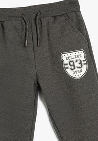Pantalones de chándal de algodón gris con cintura ajustable, bolsillo lateral y un logo en forma de escudo blanco que dice "COLLEGE 93" con estrellas. Textura suave.
