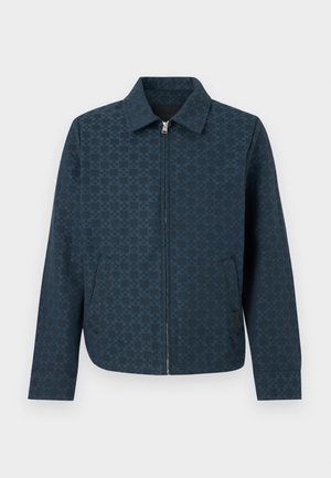 Ung mand med krøllet hår iført en mørk mønstret jakke, beige skjorte, sorte bukser med vide ben og sorte sko, stående mod en ensfarvet væg.