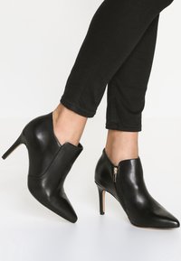 Stivaletti alla caviglia in pelle nera con punta appuntita, tacco alto e dettaglio con cerniera laterale. Finitura strutturata con un design elegante e moderno.