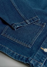 Tumši zilas džinsu jaka, kurai ir salocīta aproce, diagonāla šuve un kvadrātveida kabata ar redzamām oranžām diega šuvēm un metāla pogas detaļu.