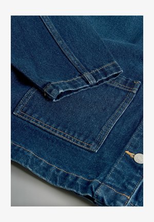 Tmavomodrá džínsová bunda s prehnutými manžetami, diagonálnym švom a štvorcovou nášivkou s viditeľným oranžovým prešívaním a kovovým gombíkom.