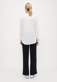 Witte blouse met lange mouwen, een rugpas en afgeronde zoom, gecombineerd met zwarte, wijde broekspijpen. Zilveren schoenen maken de outfit compleet.