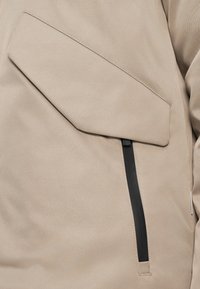 Beige puha héjú dzseki nagy, ferde zsebbel és fekete cipzárral. Az anyag sima textúrájú és minimalista dizájnnal rendelkezik.