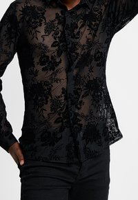 Camicia nera trasparente con bottoni, caratterizzata da ricami floreali in un motivo testurizzato, maniche lunghe e colletto classico.