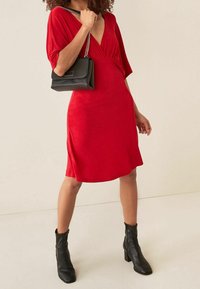 Robe rouge avec un décolleté en V profond, manches courtes et taille cintrée. Associée à un sac à main noir avec une bandoulière chaîne et des bottines noires à talons.