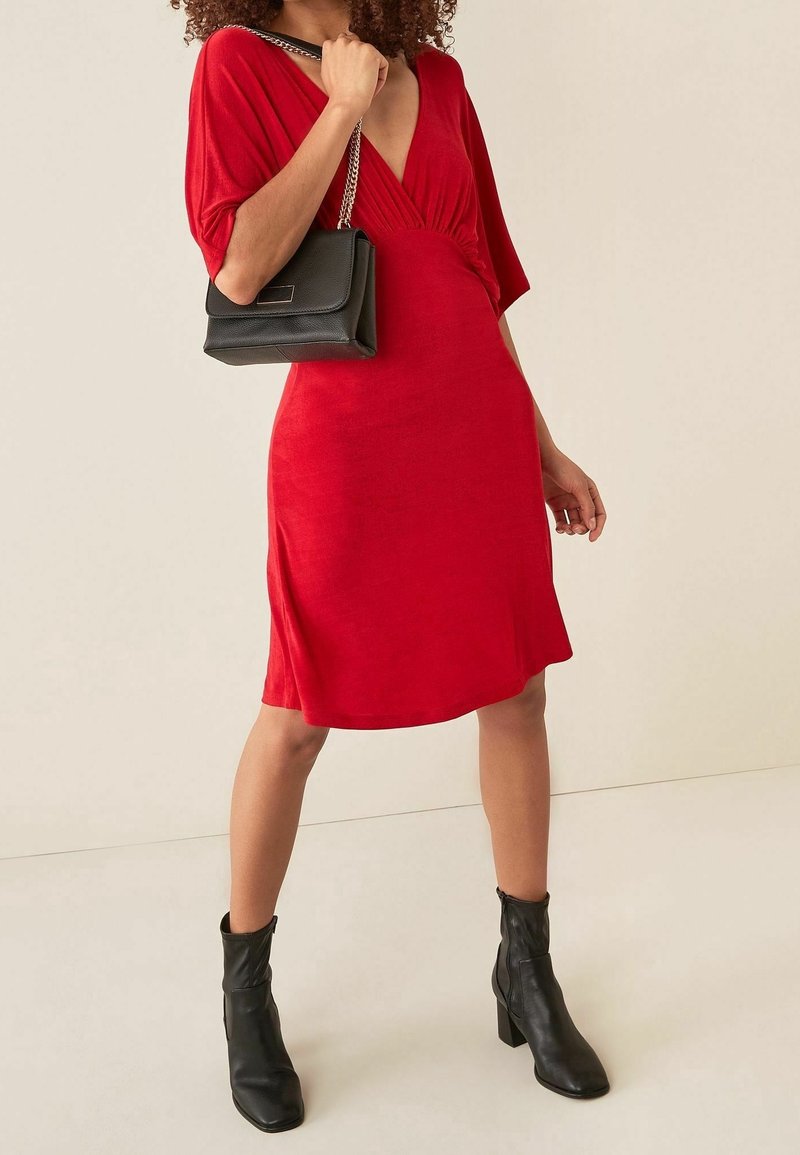 Robe rouge avec un décolleté en V profond, manches courtes et taille cintrée. Associée à un sac à main noir avec une bandoulière chaîne et des bottines noires à talons.