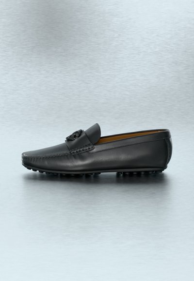 Μαύρο δερμάτινο μοκασίνι με ανάγλυφο σχέδιο στο λουρί, στρογγυλή μύτη και λάστιχο με ανάγλυφη υφή στη σόλα, απεικονίζεται σε προφίλ πάνω σε μια αντανακλαστική επιφάνεια.