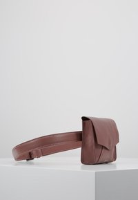 Bolso de cinturón de cuero marrón con un acabado texturizado, que presenta un cierre con solapa y una correa ajustable. Diseño minimalista sin hardware visible.