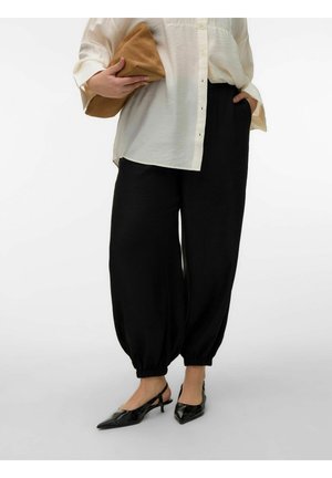 Személy laza fekete nadrágot visel rugalmas szegéllyel, krémszínű gombos inggel, fekete pántos magassarkú cipőben, és egy barna táskát tart a kezében.